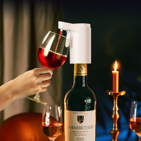 Dispensador automático de vino blanco sirviendo una copa en una mesa con una botella de vino tinto, copas adicionales y una vela encendida en un ambiente de cena.