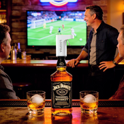 Botella de whisky con un dispensador automático Loopra en la barra de un bar, flanqueada por dos vasos con hielo redondo, con clientes y una pantalla de televisión mostrando fútbol al fondo.