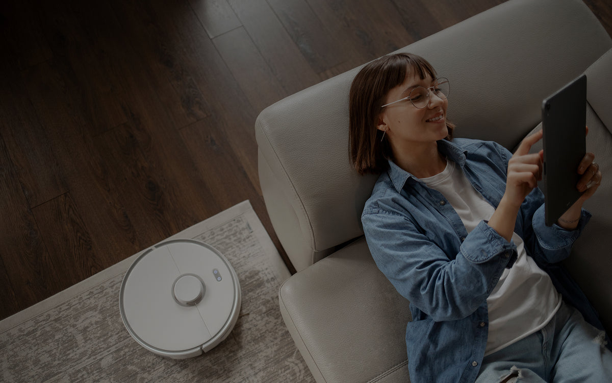 Vista aérea de una joven relajada en el sofá controlando un dispositivo desde su tableta, mientras un robot aspirador inteligente realiza la limpieza del suelo de la sala.
